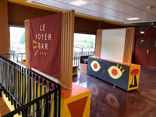 Mobilier Luxy : Conception et r&eacute;alisation d&rsquo;un mobilier original sur mesure pour le foyer bar du Luxy Cin&eacute;ma Municipal
☞ Dans le cadre du r&eacute;am&eacute;nagement du Foyer-Bar, notre collectif a travaill&eacute; avec l&rsquo;&eacute;quipe du cin&eacute;ma pour imaginer un ensemble de mobilier adapt&eacute; &agrave; l&rsquo;animation et la programmation du cin&eacute;ma. Le Foyer Bar est un endroit phare et rep&egrave;re pour les usag&egrave;r&middot;es et l&rsquo;&eacute;quipe du luxy. Il est modulable et accueille beaucoup temps forts et d&rsquo;activit&eacute;s (go&ucirc;ters, rencontres, ateliers, buffet etc.) mais c&rsquo;est aussi un espace o&ugrave; les usag&egrave;r&middot;es aiment se poser, prendre le temps; Se dessine donc un espace enfant, un espace ado, un espace d&rsquo;attente, et aussi un espace de pause et de d&eacute;gustation.Cet espace est imagin&eacute; pour que le cin&eacute;ma soit lieu de vie et de rencontre.
☞ Le r&eacute;am&eacute;nagement du Foyer Bar part d'une envie et d&rsquo;un besoin de valoriser, d&rsquo;embellir. L'objectif pour nous en tant que designers est donc d&rsquo;harmoniser, d&rsquo;optimiser et d&rsquo;inventer un ensemble de mobiliers coh&eacute;rents r&eacute;pondant &agrave; ces probl&eacute;matiques.
◼︎ Cr&eacute;ation d&rsquo;un ensemble de mobilier sur mesure &agrave; double usage :&nbsp;Des modules d&rsquo;assises avec coussins (pour augmenter le nombre d&rsquo;assises et cr&eacute;er un espace de pause) et de rangements (pour les livres, la documentation).&nbsp;Des modules d'affichages, de signal&eacute;tiques (du mobilier avec une paroi d&rsquo;affichage int&eacute;gr&eacute;e) et de rangements&nbsp; (livres, mat&eacute;riel des ateliers etc.). Pour ces deux modules nous avons r&eacute;cup&eacute;r&eacute; les tableaux aimant&eacute;s d&eacute;j&agrave; pr&eacute;sents sur site que nous avons int&eacute;gr&eacute; (dans une d&eacute;marche de r&eacute;emploi)/
◼︎A partir de la gamme color&eacute;e du Luxy et de l&rsquo;espace cin&eacute;ma, nous avons d&eacute;velopp&eacute; un univers graphique s&rsquo;inspirant de la projection(faisceau), du croisement et de la rencontre. M&ecirc;lant &eacute;l&eacute;ments graphiques g&eacute;om&eacute;triques et mall&eacute;ables, l&rsquo;identit&eacute; graphique du foyer s&rsquo;harmonise au gr&egrave;s de l&rsquo;ensemble des&nbsp;
&nbsp;